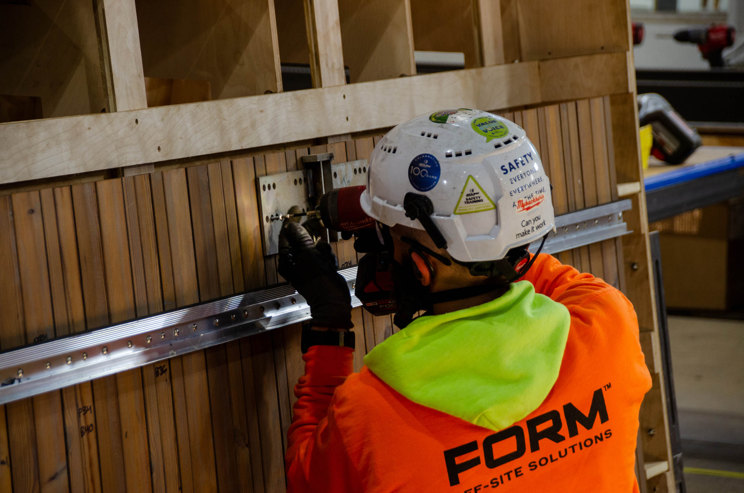 Worker in orange hi-vis jacket and white hard hat adjusting a metal bracket on a wooden installation wall.