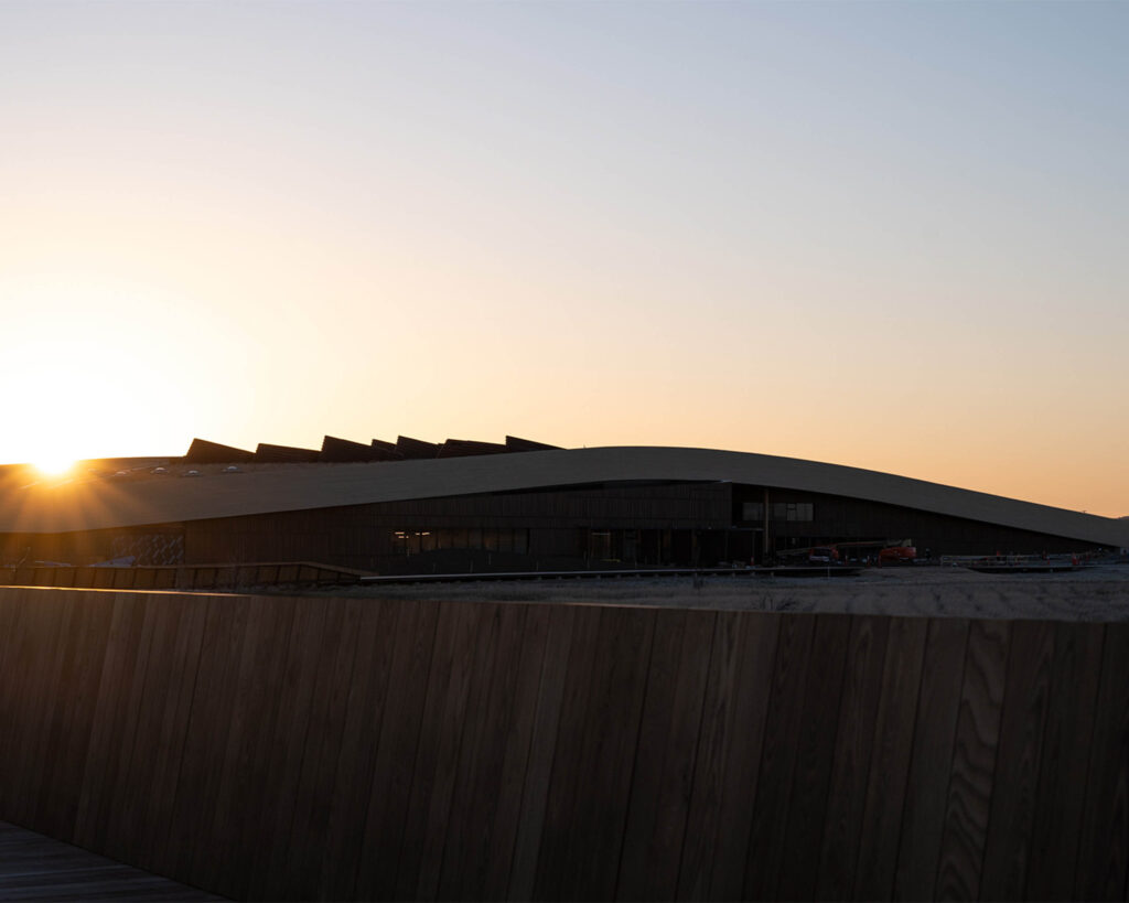 Sunset over a modern building with a slanted roof and dark wooden foreground fence in silhouette.