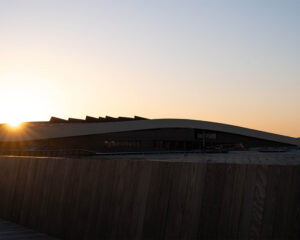 Sunset over a modern building with a slanted roof and dark wooden foreground fence in silhouette.